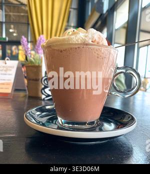 Heiße Schokolade mit Schlagsahne und bunten Streuseln serviert in einer klaren Glasschale auf einer schwarz-weißen Untertasse, mit einem gemütlichen Café Hintergrund Stockfoto