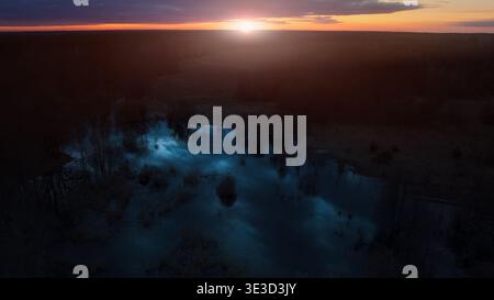 Eine Drohnenaufnahme aus der Luft fängt die ruhige Schönheit einer Dämmerungslandschaft mit einem lebendigen Sonnenuntergang ein, der den Horizont über einem dichten Wald und einer Trank beleuchtet Stockfoto