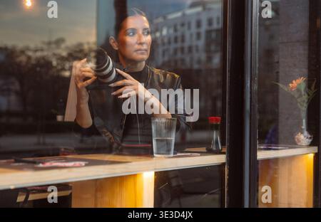 Fotografin, die Kamera im Café hält, mit städtischer Reflexion am Fenster Stockfoto