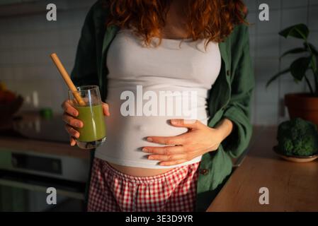 Schwangere Frau in einer Küche, die ihren Babypuffer und ein Glas belebenden grünen Smoothie hält, der eine gesunde Ernährung und ausreichende Vitamine für ein wachsendes Baby betont Stockfoto