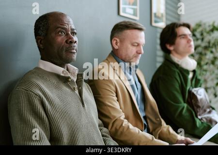 Schwarzer Mann mittleren Alters wartet mit kaukasischen Männern im Empfangsbereich der Bankfiliale Stockfoto