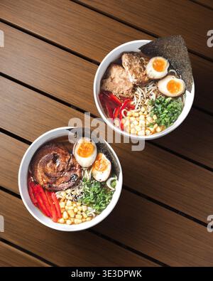 Zwei Schüsseln Ramen ruhen auf einem Holztisch. Die sanfte Beleuchtung hebt die Nudeln und Toppings hervor. Hartgekochte Eier, Gemüse und rote Paprika verleihen Farbe. Tex Stockfoto