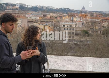 Junges Paar mit Smartphones und Blick auf das weitläufige Stadtbild von Rom, Italien, mit historischen Gebäuden und dem Petersdom in der Ferne. Stockfoto