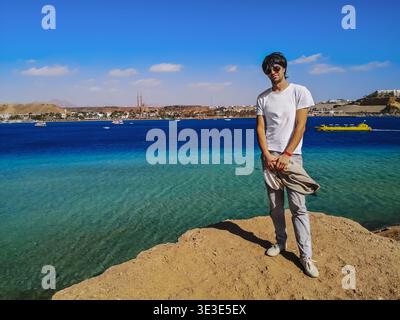 Junger dunkelhaariger Europäer posiert über einer Klippe vor dem Hintergrund des Roten Meeres in Scharm El Sheikh (Ägypten). Ein hübscher Erwachsener steht auf einem Felsen Stockfoto