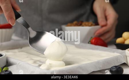 Joghurtrinde herstellen. Frau, die Joghurt mit Löffel auf das Geschirr streut, auf dem weißen Tisch, Nahaufnahme Stockfoto
