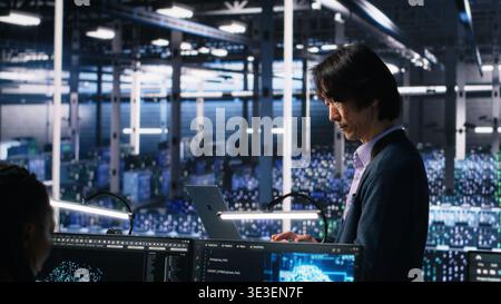Teamleiter beaufsichtigt Techniker im Serverraum mithilfe von KI zur Überprüfung der Infrastrukturdiagnose. Asiatischer Mann im Rechenzentrum, der die Leistung von Mitarbeitern mit künstlicher Intelligenz analysiert, Kamera A Stockfoto