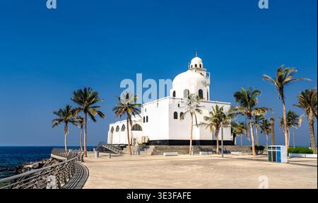 Die weiße Inselmoschee steht an der Küste des Roten Meeres an der Jeddah Corniche in Saudi-Arabien, eingerahmt von tropischen Palmen unter einem klaren blauen Himmel. Stockfoto