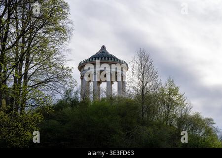 Der Monopteros, ein neoklassizistischer Säulenpavillon, steht auf einem grünen Hügel im Englischen Garten von München, umgeben von Springbäumen und hellem Himmel. Stockfoto