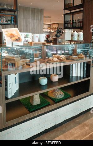 Vitrine aus Glas für verschiedene Backwaren. Die Regale sind mit dekorativen Äpfeln und grünen Matten ausgestattet. Die moderne Inneneinrichtung ist mit weißen Ziegelwänden und Holzboden ausgestattet Stockfoto