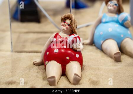 Eine lustige Figur einer molligen Dame mit rotem Badeanzug und Sonnenbrille, die mit einem Ball auf Sand sitzt. Stockfoto