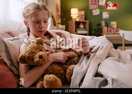 Mädchen hält Thermometer und Spielzeugbär, während sie im Bett liegt Stockfoto