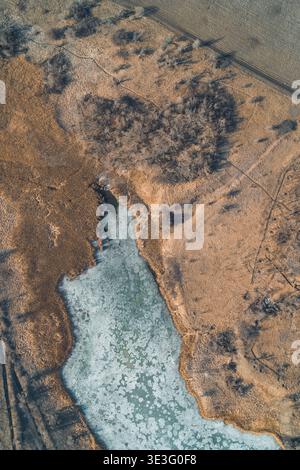 Gefrorener blauer See mit komplizierten Eisstrukturen liegt eingebettet zwischen trockenen braunen Wiesen und Blattbüschen. Diese Hochwinkellandschaft fängt den ruhigen Übergang vom Winter zum Frühling in einer ländlichen Gegend ein. Stockfoto