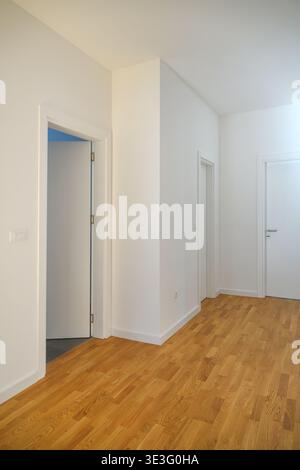 Innenansicht auf einen sonnendurchfluteten leeren Apartmentkorridor mit weißen Wänden, Eichenparkett und weißen Türen. Sauberer, minimalistischer Wohnbereich Stockfoto