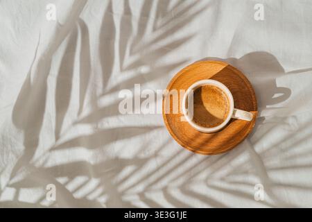 Sommertagsszene mit einer Tasse Kaffee und tropischen Palmen hinterlässt Schatten auf weißem Leinenhintergrund. Sonne und Schatten. Stockfoto