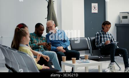 afroamerikanischer Mann und älterer Patient warten zusammen an der Krankenhausaufnahme und schauen sich die Online-Website auf dem Smartphone an. In der Empfangshalle der medizinischen Klinik sitzen verschiedene Menschen. Stockfoto