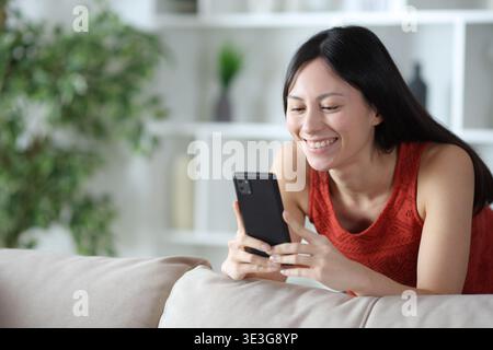 Glückliche asiatische Frau, die es genießt, das Smartphone zu benutzen, das sich zu Hause auf eine Couch stützt Stockfoto