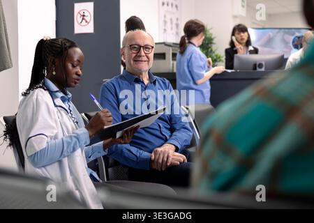 Senior man hört zu, wie afroamerikanische Ärztin im Labormantel in professionelle Gesundheitsdiskussion eingreift. Männlicher Rentner Patient schaut in die Kamera, bereit, medizinische Beratung in der Klinik Lobby zu erhalten. Stockfoto