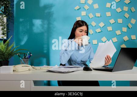 Weibliche Auditorin, die am Schreibtisch sitzt und Finanzberichte von Startup-Unternehmen analysiert. Professionelle Frau, die eine Tasse Kaffee trinkt, während sie digitale Informationen mit gedruckten Unterlagen vergleicht. Stockfoto