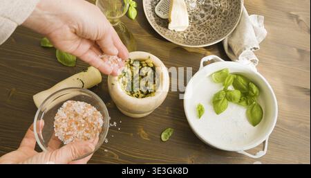 Person fügt rosafarbenes Meersalz in Marmormörtel bei, während Sie traditionelles Genoveses Pesto herstellen. Nahaufnahme der Hände mit einem kleinen Holzlöffel zum Hinzufügen von Himalaya-Salz Stockfoto