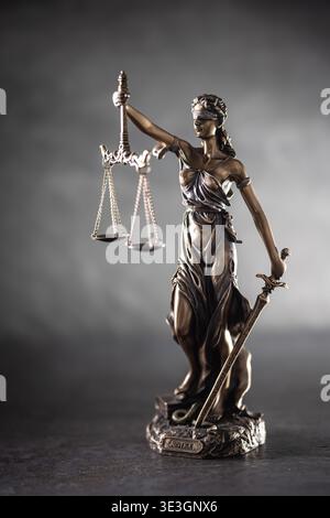 Eine Bronzestatue der Gerechtigkeit steht vor einem dunklen, strukturierten Hintergrund mit dramatischer Seitenbeleuchtung. Die Figur hält Waagen, die Gerechtigkeit in einem symbolisieren Stockfoto