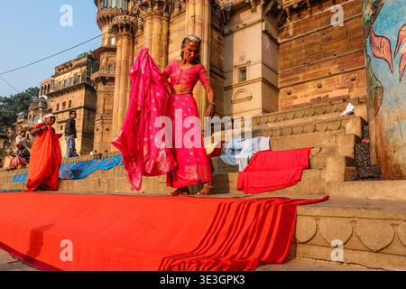 Eine Frau in einem rosafarbenen Sari breitet einen langen roten Stoff aus, um auf den Stufen des Munshi Ghat in Varanasi zu trocknen, mit historischen Sandsteinpalästen darüber. Stockfoto