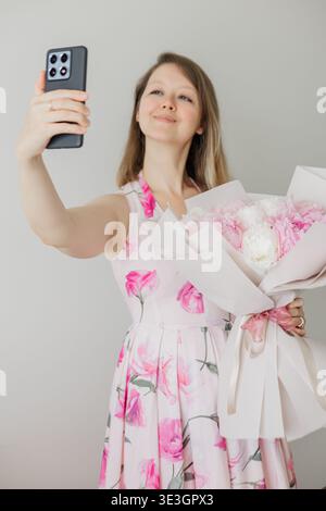 Eine junge Frau, die Selfie nimmt und einen Strauß rosa Pfingstrosen hält Stockfoto