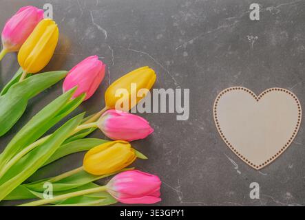 Leuchtende gelbe und rosafarbene Tulpen auf dunklem Marmor mit einem hölzernen Herzzeichen. Romantisches Stillleben im Frühling mit stimmungsvoller Beleuchtung und hochwertigen Texturen Stockfoto