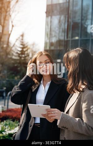 Zwei Frauen diskutieren über die Arbeit, während sie ein Tablet halten. Sie stehen in einem Stadtgebiet mit modernen Gebäuden und Grün um sie herum, enj Stockfoto