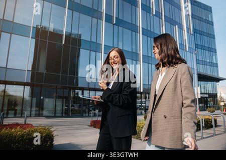 Zwei Frauen laufen zusammen vor einem großen Bürogebäude in der Stadt. Sie sind in Geschäftskleidung gekleidet und führen ein Gespräch, während man hält Stockfoto