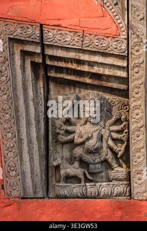 Diese Nahaufnahme im Shree Annapurna Mandir in Varanasi zeigt eine mehrarmige Gottheit, die in eine dunkle Holztafel geschnitzt ist, die in eine hellrote Mauerwand eingebettet ist. Stockfoto