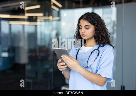 Junge Ärztin trägt Peelings und Stethoskop mit einem digitalen Tablet und verwaltet Patientendaten in einer modernen Klinik oder Krankenhaus-Umgebung mit fortschrittlicher Gesundheitstechnologie Stockfoto