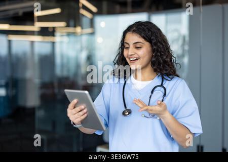 Junge Ärztin oder Krankenschwester in blau schrubbt mit einem digitalen Tablet für eine virtuelle Patientenberatung, kommuniziert und bewegt fröhlich während eines Remote-Arzttermins Stockfoto