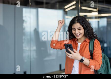 Junge lächelnde Frau, die Erfolge und Erfolge feiert, während sie ihr Smartphone überprüft und in einer modernen Büro- oder Bildungsumgebung triumphiert Stockfoto