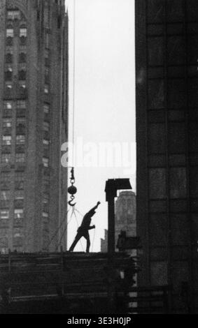 Dieses düstere Straßenfoto, das wahrscheinlich von Angelo Rizzuto in den 1940er Jahren aufgenommen wurde, bildet einen Bauarbeiter inmitten der hohen Wolkenkratzer von New York City dramatisch. Der Arbeiter scheint Baumaterial zu führen, das von einem Kran hochgehoben wird, was das kontinuierliche Wachstum der Stadt und die Arbeit hervorhebt, die mit ihrer beeindruckenden Architektur verbunden ist. Stockfoto