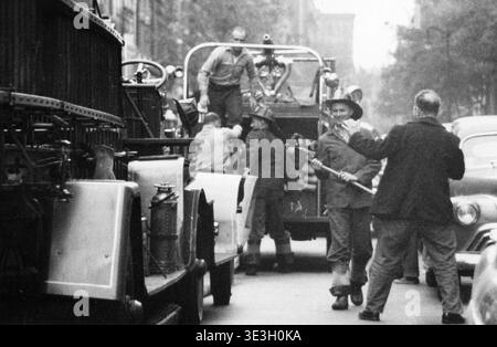 Dieses alte Straßenfoto, wahrscheinlich aus den 1940er Jahren von Angelo Rizzuto, zeigt einen ehrlichen Moment der Feuerwehrleute in Greenwich Village, New York City, nach Abschluss eines Jobs. Ein Feuerwehrmann in einem Schutzhelm lächelt, während er mit einer anderen Person interagiert, während andere mit Ausrüstung rund um ein Feuerwehrfahrzeug beschäftigt sind und den Alltag und die gelegentlichen Interaktionen dieser Zeit verkörpern. Stockfoto
