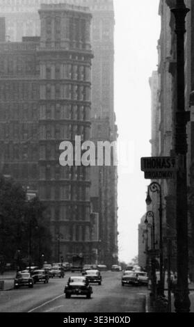 Ein stimmungsvolles Schwarz-weiß-Straßenfoto von Angelo Rizzuto, wahrscheinlich aus den 1950er Jahren, das einen Blick auf den Broadway in New York City mit einem Straßenschild für „THOMAS ST + BROADWAY“ zeigt. Das Bild zeigt Oldtimer, die die Straße entlang fahren, flankiert von hoher, klassischer New Yorker Architektur unter einem bedeckten Himmel. Stockfoto