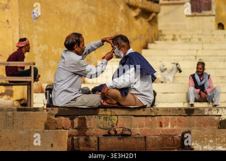 Ein Straßenfriseur rasiert den Bart eines Kunden, während beide im Kreuz auf einem Steinvorsprung in einem Varanasi Ghetto am Ufer des Ganges sitzen. Stockfoto