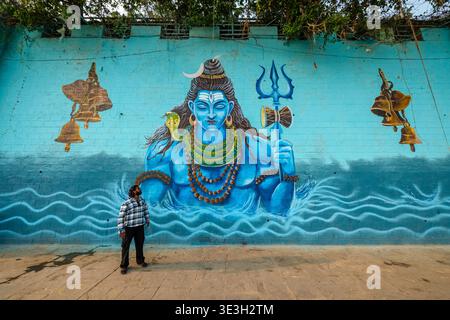 Ein Mann steht vor einem großen, leuchtend blauen Wandbild von Lord Shiva, gemalt auf einer hohen Wand in einem Varanasi Ghetto am Fluss Ganges. Stockfoto