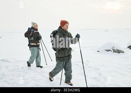 Kaukasische Männer und Frauen mittleren Alters wandern mit Wanderstöcken durch verschneite Landschaft, beide tragen Rucksäcke und Winterhüte und gehen Seite an Seite in der Winterwildnis Stockfoto
