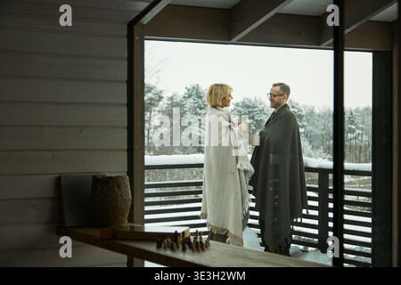 Kaukasische Frau und Mann mittleren Alters, die auf dem Balkon stehen, in Decken gehüllt, mit Bechern, die sich mit verschneiten Wäldern unterhalten, die im Hintergrund sichtbar sind Stockfoto