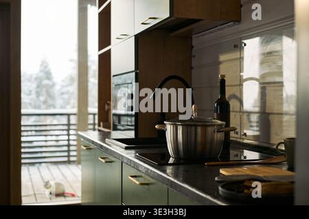 Moderne Küchenarbeitsfläche mit Edelstahltopf mit Deckel auf Induktionsherd, Weinflasche in der Nähe, verschwommener Balkonblick mit Katze draußen im Hintergrund Stockfoto
