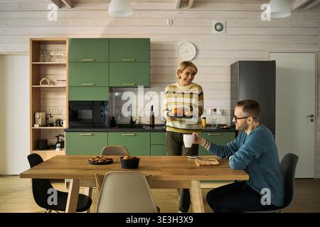 Kaukasische Frau mittleren Alters, die in der modernen Küche steht, einen Teller hält, lächelt einen kaukasischen Mann mittleren Alters an, der am Holztisch sitzt und Kaffee trinkt und sie ansieht Stockfoto