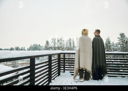 Kaukasische Erwachsene Frau und Mann stehen auf einem schneebedeckten Balkon, in Decken gewickelt, einander gegenüber und blicken auf die Winterwaldlandschaft im Hintergrund Stockfoto