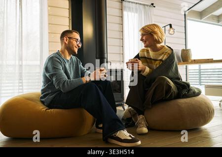 Kaukasischer Erwachsener Mann und Frau sitzen auf Fußbodenkissen gegenüber, lächeln und halten Tassen in der Hand und führen entspannte Gespräche in einem modernen Wohnzimmer Stockfoto