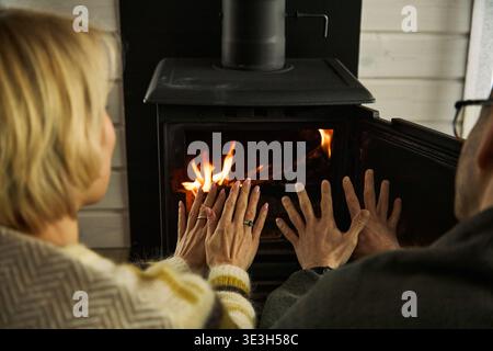 Kaukasische Frau mittleren Alters und kaukasischer Mann mittleren Alters wärmen Hände zusammen vor dem Holzofen, sitzen dicht und mit Blick auf den offenen Kamin, genießen Wärme drinnen Stockfoto