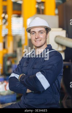 Porträt des kaukasischen Ingenieurs männlich, glücklicher Arbeiter junger Mann, der zuversichtlich den Arm überkreuzt und lächelnd in der Fabrik der Arbeitsplatzindustrie mit Sicherheitsuniform steht Stockfoto