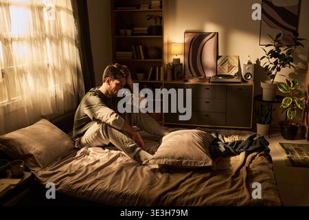 Junger Mann, der auf dem Bett sitzt und im Schlafzimmer gestresst wirkt Stockfoto
