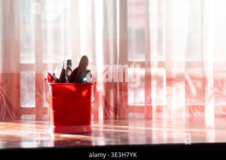 Weitwinkelaufnahme von Besteck in einem roten Behälter auf einer Holzoberfläche, mit hellem natürlichem Licht aus einem Fenster mit viel Kopierraum. Stockfoto