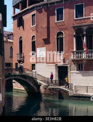 Blick auf eine Brücke, die über einen Kanal führt und die Terrakotta-Gebäude reflektiert, wo die Menschen entlang der Straße laufen, Venedig, Venetien, Italien. Stockfoto