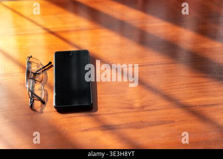 Hochwinkelansicht eines Mobiltelefons und einer Brille auf einer Holzoberfläche, mit langen Schatten aus einem Fenster in warmer Morgensonne. Stockfoto